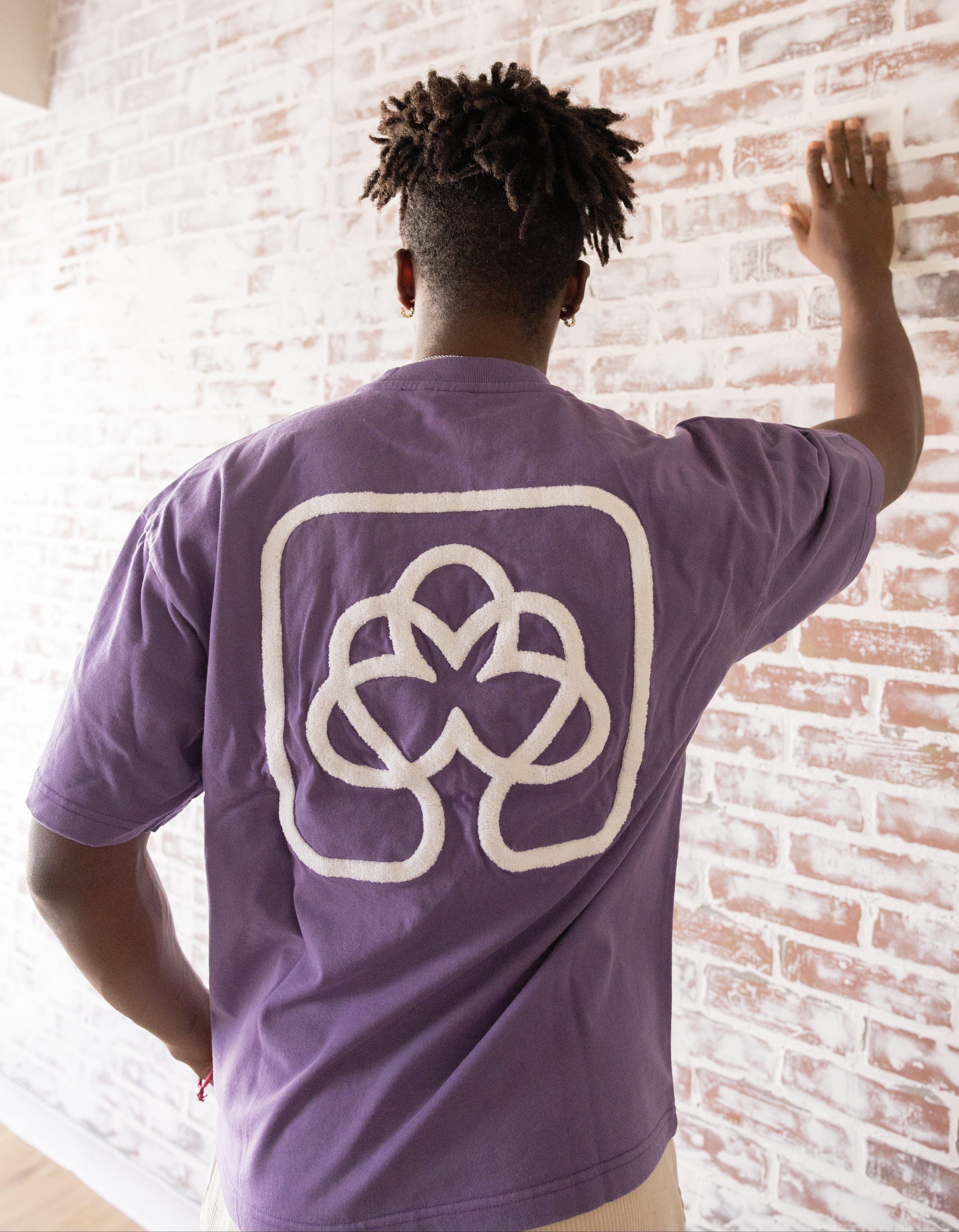 Person wearing a purple t-shirt with a white logo against a brick wall.